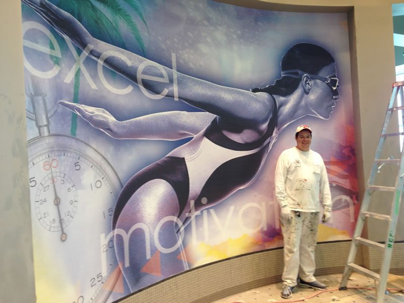 Wayne standing in front of a large-scale mural he painted across a commercial wall.